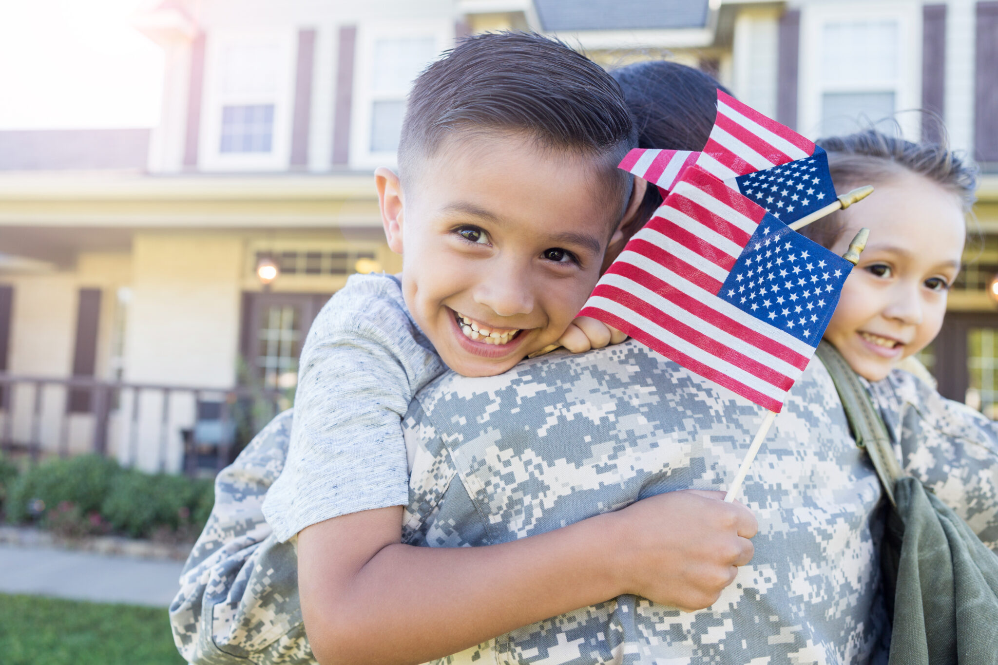 oung, Hispanic boy smiles as he holds a flag in each hand and smiles. He and his sister hug his mother in uniform who has just arrived home.